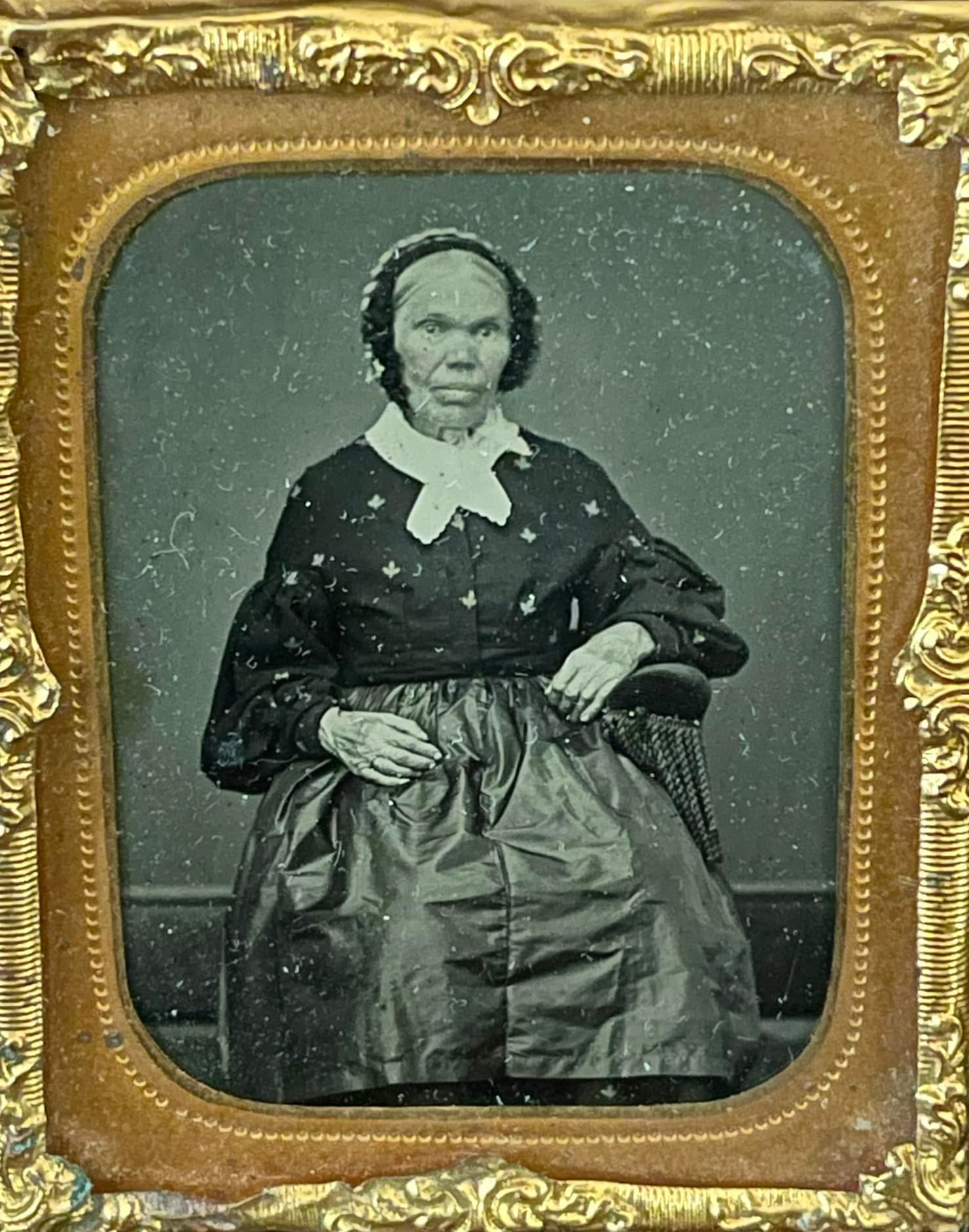 A black-and-white photograph—perhaps a daguerreotype—of an older African American woman. She is seated and wears a bonnet, a dark shirt with a large white collar, and a shiny skirt. The photograph is set in a tarnished, golden frame with a mustard-yellow mat.