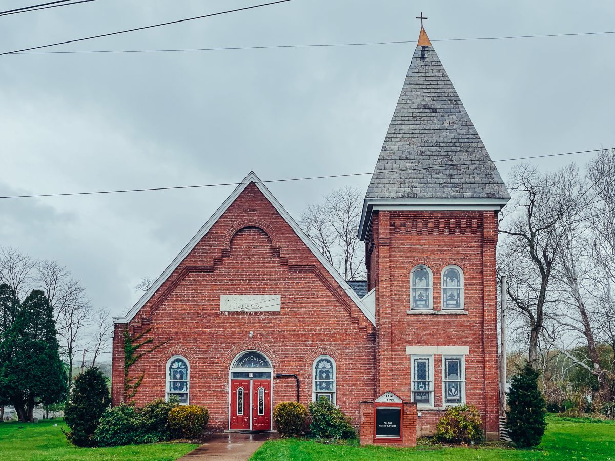 A contemporary photograph of a red brick church set against a grey sky. Electric-green grass surrounds the building, which is adorned with an equally bright red door, stained-glass windows, and a steeple topped by a cross. There is a stone marker on the church that reads “A. M. E. Church 1902.”