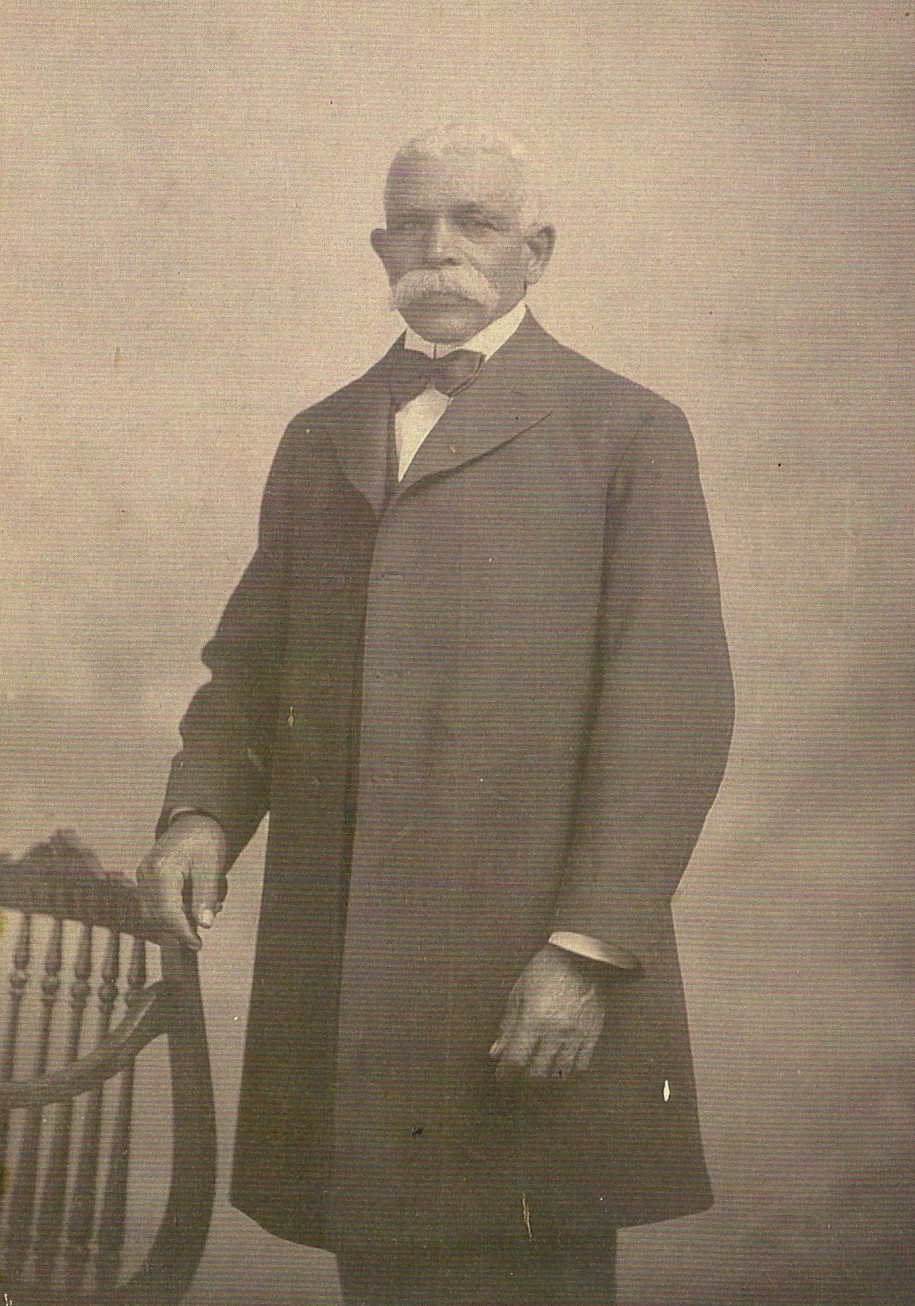 A photograph of an older African American man. He is standing tall with white hair and a large white mustache, his right hand lightly touching a wooden chair. He is dressed in a white collared shirt with a dark bow tie and long overcoat that appears to be the same fabric as the tie.