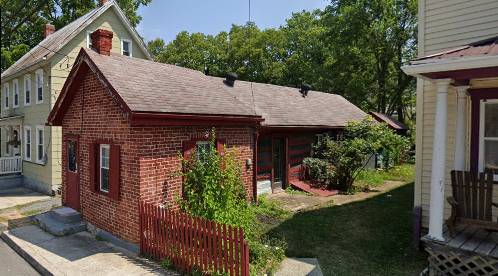 A contemporary photograph of a modest, single-story red brick home. The trim, including the door, window shutters, and picket fence, is a slightly darker shade of red. Bright green shrubs and trees grow in the side yard. The roof is made of asphalt shingles, faded by the sun. 